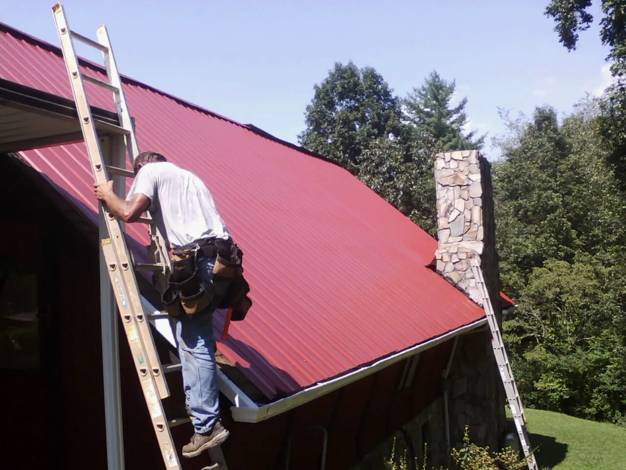 Roofer on a ladder beside a red metal roof and stone chimney — Soffit & Fascia Repair by Roofing Brothers in Galena